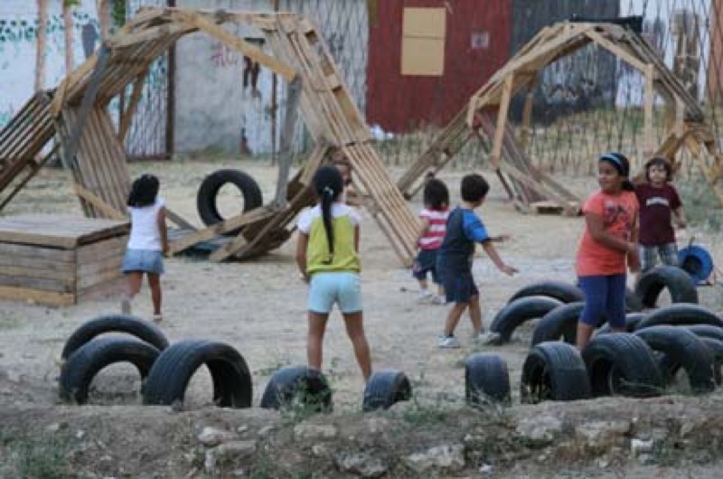 Zona de juego de niños con compostera, elementos representativos y vallado al fondo