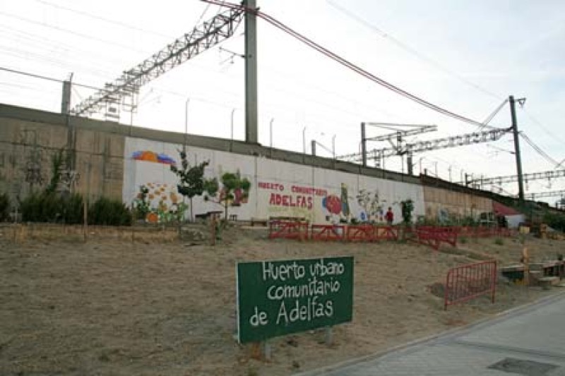 Adelfas. Vista general de la parcela desde la calle con el huerto, el mural y las vías del tren al fondo